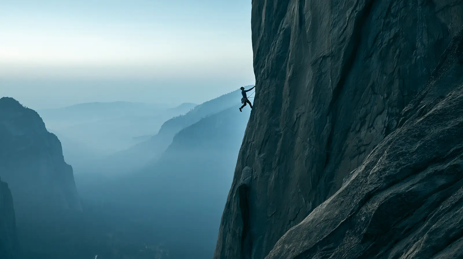 A climber hangs from a sheer granite wall above a misty valley.