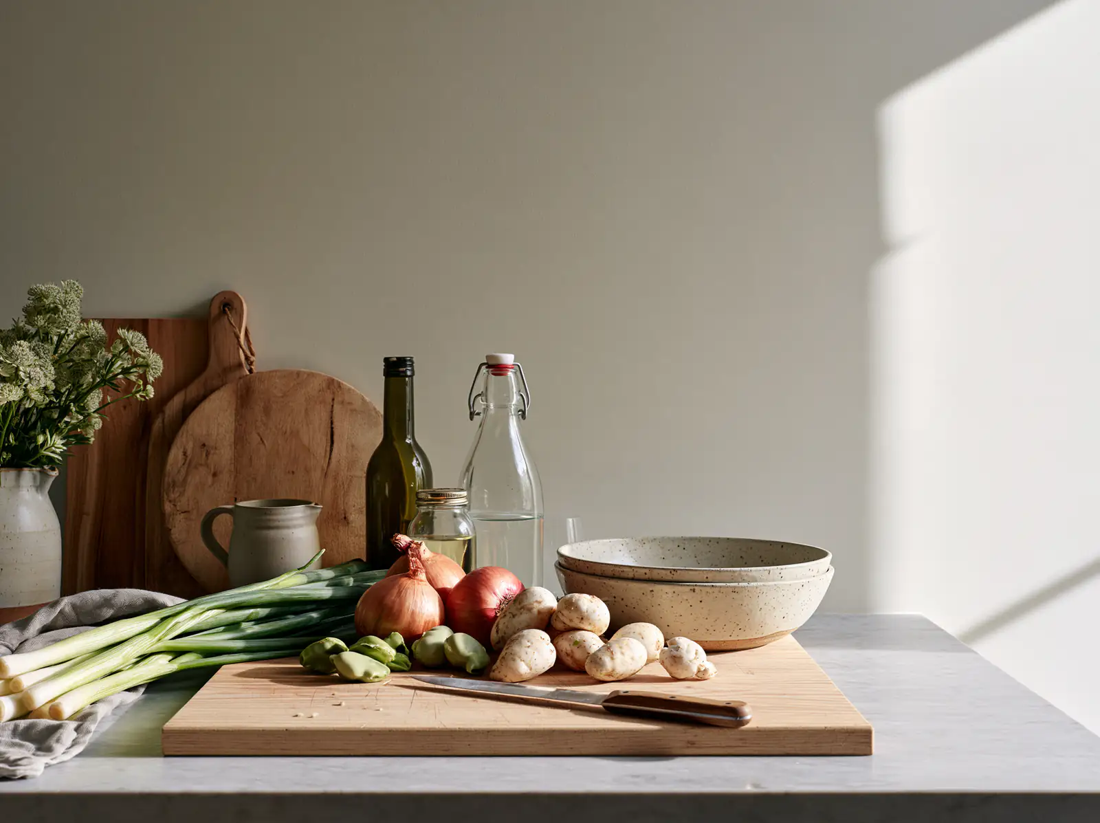 Leeks, onions, potatoes and a knife on a wooden board in low morning sun.