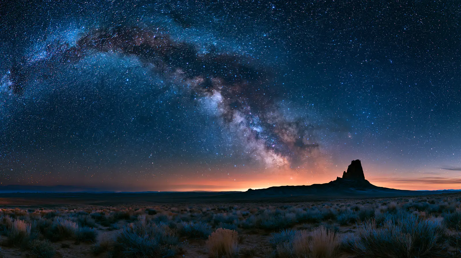 The Milky Way arches over a desert with a lone rock tower on the horizon.