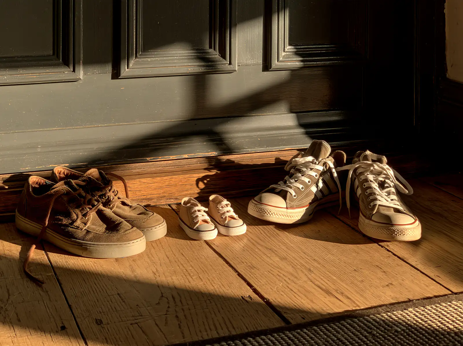 Two pairs of worn adult trainers by a door with a tiny pair of baby shoes between them.