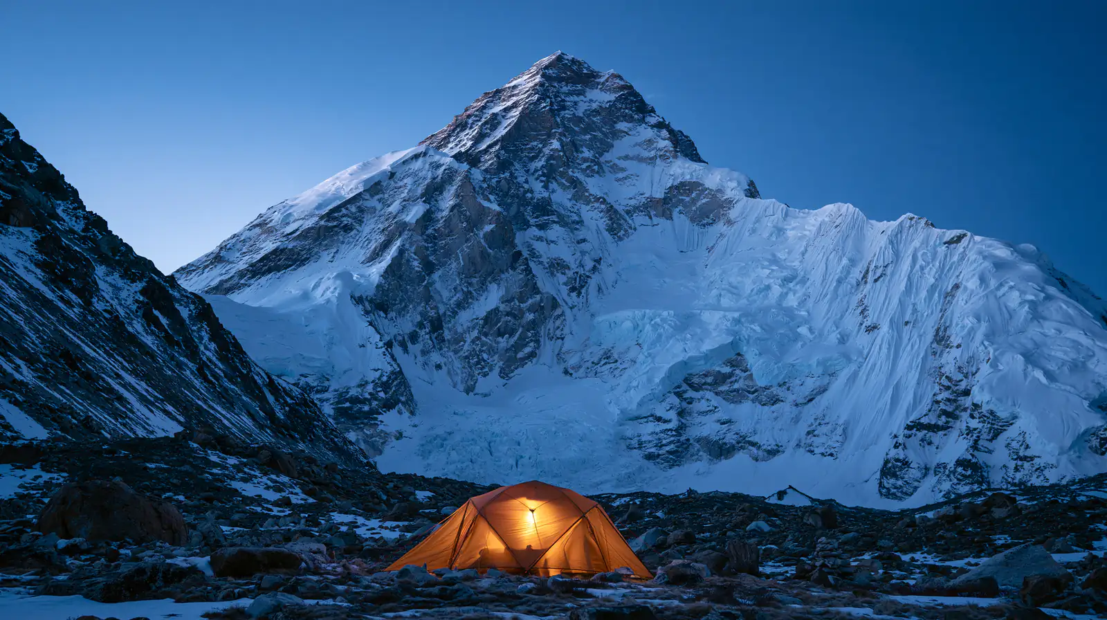 A lit orange tent below a vast snow-covered mountain at dusk.